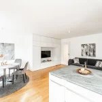 Blick von der Küche in ein helles, modernes Wohnzimmer: linker Teil zeigt einen runden Esstisch mit Stühlen, rechts ein Sofa vor Gemälden. Weiße Einbaumöbel inklusive TV. Holzboden und abgehängte Decken mit integrierter Beleuchtung.