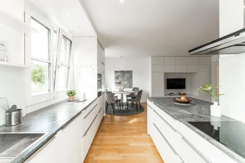 Moderne Küche mit Essbereich und Blick ins Grüne Das Bild zeigt eine moderne, helle Küche mit weißen Schränken und dunkler Arbeitsplatte. Links im Bild ist ein Fenster. Durch die offene Raumgestaltung sind ein Essbereich mit rundem Teppich und grauen Stühlen sowie ein Wohnzimmer mit Fernseher erkennbar.