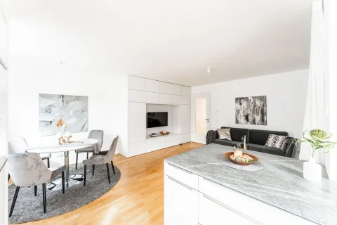 Modernes Wohngefühl: Offener Blick von der Küche Blick von der Küche in ein helles, modernes Wohnzimmer: linker Teil zeigt einen runden Esstisch mit Stühlen, rechts ein Sofa vor Gemälden. Weiße Einbaumöbel inklusive TV. Holzboden und abgehängte Decken mit integrierter Beleuchtung.