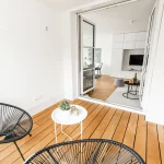 Das Bild zeigt eine stilvoll eingerichtete Loggia mit Holzboden. Zwei moderne schwarze Drahtstühle und ein kleiner weißer Tisch stehen im Vordergrund. Die geöffnete Glas-Schiebetür gibt den Blick in ein helles Wohnzimmer mit weißer Einrichtung frei.