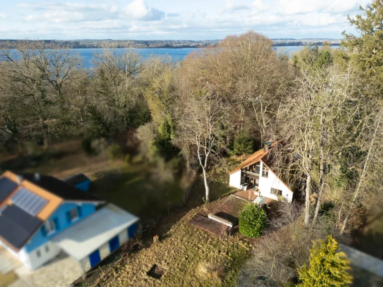 Luftaufnahme eines Gebäudekomplexes am Ufer des Ammersees. Vorne links ein Haus mit blauem Dach und Solarpaneelen. Dahinter ein kleineres, weißes Haus mit braunem Dach. Viel Grünfläche und Bäume. Im Hintergrund der Ammersee und noch mehr Bäume.