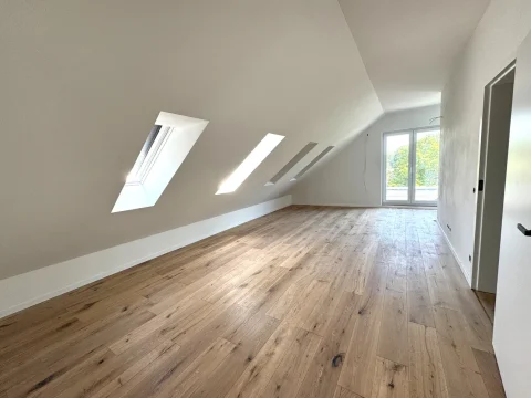 Das Bild zeigt ein leeres Dachgeschoss mit schrägen Wänden. Es sind vier Dachfenster und ein großes Fenster an der Stirnseite sichtbar. Der Boden ist mit hellbraunem Holzparkett ausgelegt. Die Wände sind weiß gestrichen.