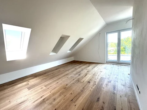 Das Bild zeigt ein helles Dachstudio mit schrägen Wänden. Es sind drei Dachfenster und eine große Fenstertür zu sehen, die viel Licht hereinlassen. Der Boden ist mit Holzdielen ausgelegt. Die Wände sind weiß gestrichen.
