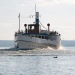 Ein weiß-blauer Dampfer fährt auf dem Ammersee. Passagiere stehen auf dem Deck. Im Hintergrund sind bewaldete Hügel und ein klarer Himmel. Wellen brechen am Bug des Schiffes.