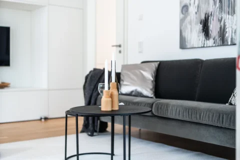 Moderne Wohnzimmerästhetik: Eleganz in Weiß und Grau Ein Wohnzimmer mit modernem Interieur: rechts graues Sofa mit Kissen, links kleiner runder Tisch mit Kerzenhaltern und einer Glühbirne. Hintergrund zeigt weiße Regale, ein Gemälde und einen Fernseher.