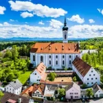 Panoramablick über Dießen am Ammersee: im Vordergrund eine große Kirche mit Turmuhr und grünem Vorplatz, dahinter dicht bebautes Dorf mit roten Dächern, dahinter Felder und die Alpen. Himmel ist klar mit einigen Wolken.