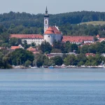 Das Bild zeigt die Stadt Diessen am Ammersee mit dem markanten Marienmünster. Die Kirche mit weißer Fassade und rotem Dach steht im Vordergrund, dahinter das Ufer und bewaldete Hügel. Im Vordergrund ist der Ammersee mit mehreren Booten.