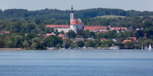 Das Bild zeigt die Stadt Diessen am Ammersee mit dem markanten Marienmünster. Die Kirche mit weißer Fassade und rotem Dach steht im Vordergrund, dahinter das Ufer und bewaldete Hügel. Im Vordergrund ist der Ammersee mit mehreren Booten.