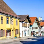 Bunte Häuserreihen mit traditioneller Architektur entlang einer Straße. Gelbes, grünes und weißes Gebäude, teilweise mit Läden im Erdgeschoss, in Diessen am Ammersee. Blauer Himmel und fast kahler Baum ergänzen die Szene.