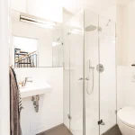 Ein modernes Badezimmer mit einer gläsernen Duschkabine, rechts davon eine Toilette und ein an der Wand montiertes Waschbecken links. Es sind Handtücher sichtbar; die Raumbeleuchtung ist hell. Im Hintergrund sieht man eine Treppe.