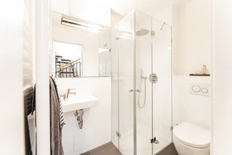 Moderne Dusche in hellem Badezimmerdesign Ein modernes Badezimmer mit einer gläsernen Duschkabine, rechts davon eine Toilette und ein an der Wand montiertes Waschbecken links. Es sind Handtücher sichtbar; die Raumbeleuchtung ist hell. Im Hintergrund sieht man eine Treppe.