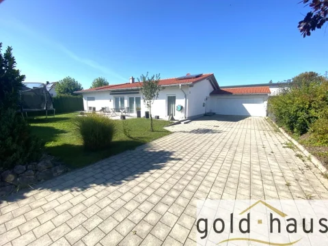 Ein weißes Einfamilienhaus mit flachem Dach und angebauter Doppelgarage. Eine gepflasterte Einfahrt führt zum Haus. Im Vorgarten sind Grünflächen, Büsche und eine Terrasse mit Sitzecke. Hinter dem Haus ist eine Trampoline sichtbar.