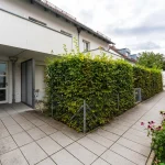 Das Bild zeigt den Eingangsbereich eines weißen Hauses in Obermenzing. Man sieht eine Glastür mit der Hausnummer 76a auf einem Schild. Neben der Tür wächst eine grüne Hecke, und im Vordergrund ist ein gepflasterter Weg sichtbar.