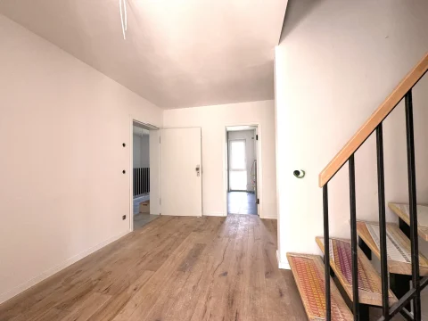 Das Bild zeigt einen Eingangsbereich in einem Haus mit Holzböden, weißen Wänden und einer rechtsseitigen Treppe mit schwarzem Geländer und Holzstufen. Links sind zwei weiße Türen und am Ende des Flurs eine weitere Tür sichtbar.