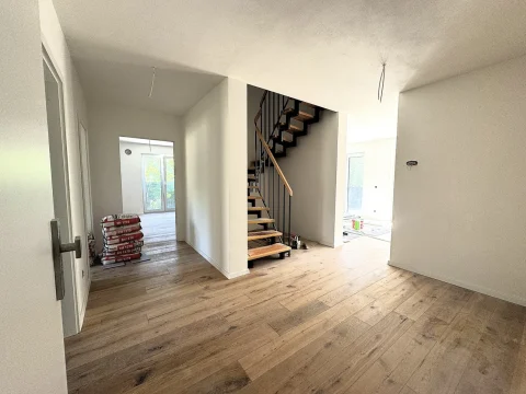 Das Foto zeigt den Eingangsbereich einer Wohnung im Obergeschoss. Zu sehen sind eine geöffnete Türe links, eine moderne Treppe rechts und Packungen mit Baumaterialien auf dem hölzernen Boden. Ein weiterer Raum ist durch eine Tür geradeaus sichtbar.
