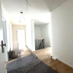 Das Bild zeigt einen langen Flur in einer Wohnung, wahrscheinlich im Eingangsbereich. Links ist eine Tür sichtbar, rechts führt der Flur zu einer Treppe mit Geländer. Der Boden kombiniert Fliesen im Eingang und Holzdielen weiter hinten. Hängendes Kabel einer Lampe sichtbar.