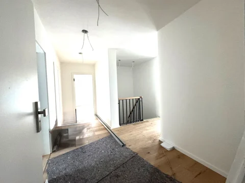 Das Bild zeigt einen langen Flur in einer Wohnung, wahrscheinlich im Eingangsbereich. Links ist eine Tür sichtbar, rechts führt der Flur zu einer Treppe mit Geländer. Der Boden kombiniert Fliesen im Eingang und Holzdielen weiter hinten. Hängendes Kabel einer Lampe sichtbar.
