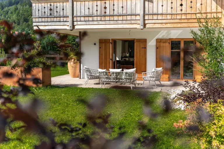 Erdgeschosswohnung mit Terrasse, die durch eine Hecke leicht verdeckt ist. Gartenmöbel-Set vor großen Glastüren, die zum hellen Wohnbereich führen. Holzverkleidung am Balkon darüber, malerischer Grünblick und Bergkulisse. Ideal für "Wohnung am Schliersee kaufen".