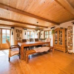 Das Bild zeigt einen rustikalen Essbereich im Landhausstil am Tegernsee mit einem großen Holztisch, umgeben von Holzbänken. Der Raum hat sichtbare Holzbalken, eine geschmückte Vitrine und liebevoll dekorierte Fenster. Licht scheint durch große Fenster.