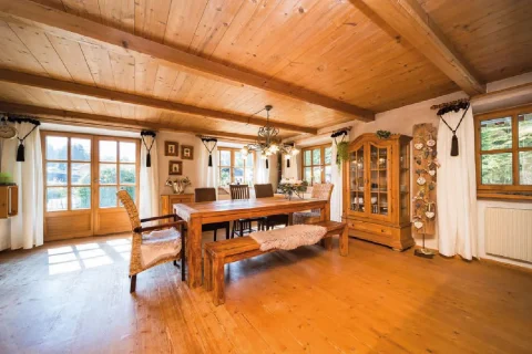 Essbereich im Landhausstil am Tegernsee Das Bild zeigt einen rustikalen Essbereich im Landhausstil am Tegernsee mit einem großen Holztisch, umgeben von Holzbänken. Der Raum hat sichtbare Holzbalken, eine geschmückte Vitrine und liebevoll dekorierte Fenster. Licht scheint durch große Fenster.