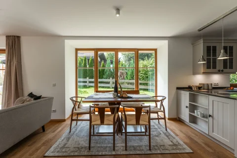 Moderne Essküche mit großem Fensterblick ins Grüne. Esstisch aus Holz, umgeben von Stühlen, teilt den Raum mit einer weißen Küchenzeile und eingebautem Kochfeld und Schrank. Elegant, geräumig mit Zugang zur Terrasse.