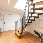 Helles, modernes Wohnzimmer mit einer schwebenden Holztreppe aus Eiche, schwarzen Metallschienen, einem kleinen Arbeitsbereich mit Tisch und Stuhl, weißen Einbauschränken und einer weißen Tür im Flur. Der Boden ist aus Parkett.