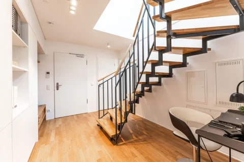 Moderner Flur mit eleganter Holztreppe und Designermöbeln Helles, modernes Wohnzimmer mit einer schwebenden Holztreppe aus Eiche, schwarzen Metallschienen, einem kleinen Arbeitsbereich mit Tisch und Stuhl, weißen Einbauschränken und einer weißen Tür im Flur. Der Boden ist aus Parkett.