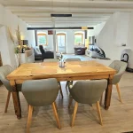 Ein rustikaler Holztisch mit vier grauen Stühlen prägt das Esszimmer. Im Hintergrund siehst du das Wohnzimmer mit einem roten Sessel, einer Couch und einem Fernseher. Die offene Raumgestaltung verbindet beide Bereiche nahtlos.