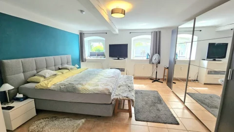 Ein helles Gästezimmer mit einem großen Bett, blau-türkiser Wand, zwei Fernsehern, einer Standventilator und Spiegel. Es gibt auch zwei Fenster, Teppiche und eine Deckenlampe. Der Raum wirkt modern und einladend.