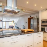 Moderne, offene Küche mit weißen Schränken, Granitarbeitsplatte und hochwertigen Geräten wie Kühlschrank und Ofen. Offener Übergang zum Wohnbereich mit Sofa und Sesseln. Stilvolle Beleuchtung und Holzbalken an der Decke.