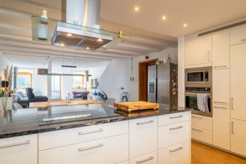 Moderne, offene Küche mit weißen Schränken, Granitarbeitsplatte und hochwertigen Geräten wie Kühlschrank und Ofen. Offener Übergang zum Wohnbereich mit Sofa und Sesseln. Stilvolle Beleuchtung und Holzbalken an der Decke.