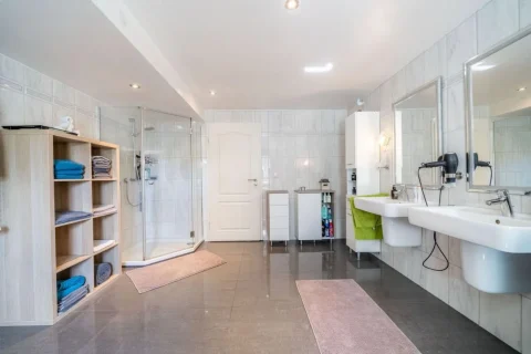 Das Bild zeigt ein großes Bad in einem modernen Stil: Es gibt zwei Waschbecken mit Spiegeln, eine geräumige Duschkabine, ein Regal mit Handtüchern und zwei kleine Schränke. Die Farbgebung ist in Weiß- und Grautönen gehalten.