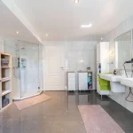 Ein großes, helles Badezimmer mit grauen Fliesen, zwei Waschbecken, großen Spiegeln, einer Glasduschkabine und einem weißen Regal voller Handtücher.