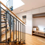 Moderne Eingangshalle mit Holztreppe, schwarzem Metallgeländer, einer eingebauten weißen Schrankwand und einer Garderobe rechts, bestehend aus Holzbänken und Fächern. Ein lichtdurchfluteter Raum mit Holzboden.
