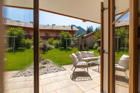 Die Aufnahme zeigt eine Terrasse an einer Wohnung am Schliersee. Man sieht eine gemütliche Sitzgruppe mit Kissen, einen gepflegten Rasen, Blumen und einen Kinderspielplatz. Im Hintergrund sind Berge und Holzhäuser.