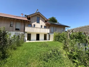 Das Bild zeigt eine ländliche Gartenansicht mit einem modernen, zweistöckigen Haus, dessen Fassade aus Holz und Putz besteht. Im Vordergrund ist eine dichte Wiese mit hohem Gras und Wildblumen, neben dem Haus steht ein alter Holzbalken. Der Himmel ist klar und blau.