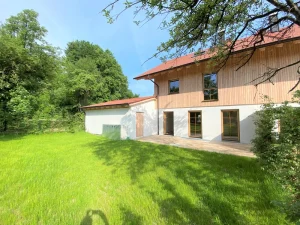 Das Bild zeigt ein Doppelhaus in einer grünen Gartenlandschaft. Die Vorderseite des Hauses ist teils aus Holz, teils verputzt, mit mehreren Fenstern und Glastüren, die auf eine Terrasse führen. Links ist eine kleinere Garage sichtbar. Der Himmel ist blau.