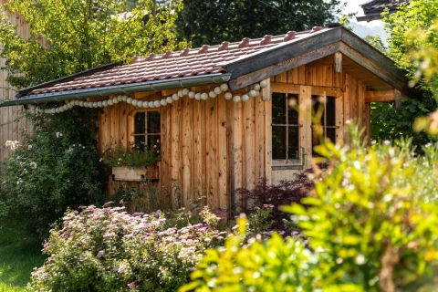 Ein charmantes Gartenhaus in Schliersee. Holzbau mit Ziegeldach, umgeben von üppigen Blumen und Pflanzen. Fenster mit Vorhängen und eine Tür bieten einen einladenden Blick. Lampions schmücken den Dachvorsprung.