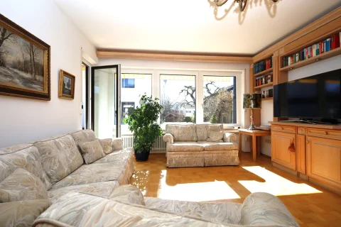 Ein gemütliches Wohnzimmer mit chremefarbenen Sofas, Holzmöbeln und Bücherregalen. Große Fenster lassen viel Licht herein und bieten einen Blick nach draußen. An den Wänden hängen Gemälde, und eine Pflanze steht im Raum.