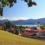 Das Bild zeigt eine Panoramaansicht von Gmund am Tegernsee. Zu sehen sind eine Kirche mit einem charakteristischen weißen Turm, umgebende Gebäude und Wiesen. Im Hintergrund erstreckt sich der Tegernsee und die bayerischen Alpen.