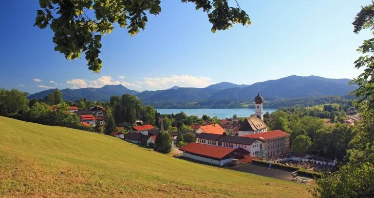 Das Bild zeigt eine Panoramaansicht von Gmund am Tegernsee. Zu sehen sind eine Kirche mit einem charakteristischen weißen Turm, umgebende Gebäude und Wiesen. Im Hintergrund erstreckt sich der Tegernsee und die bayerischen Alpen.