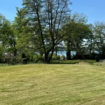 Das Bild zeigt ein gepflegtes Grundstück am Ammersee mit grünem Rasen, verschiedenen Laubbäumen und Blick auf den See. Ideal für Käufer, die Natur und Wasser schätzen.
