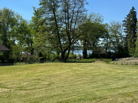 Das Bild zeigt ein gepflegtes Grundstück am Ammersee mit grünem Rasen, verschiedenen Laubbäumen und Blick auf den See. Ideal für Käufer, die Natur und Wasser schätzen.
