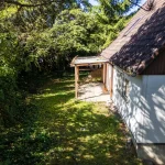 Das Bild zeigt ein teilweise schattiges Grundstück mit einem älteren Gebäude, davon sind Wände aus weißem Putz und ein dunkles Schindeldach zu sehen. Eine Holzlaube steht neben dem Haus, eingebettet in eine naturbelassene Umgebung mit Bäumen und Sträuchern.