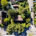 Luftaufnahme einer Straße mit einzelnen Häusern und einem freien Grundstück, möglicherweise in München. Dichte Begrünung umgibt die Gebäude und Autos parken am Straßenrand. Ideal für "Grundstück zu verkaufen" Interessenten.