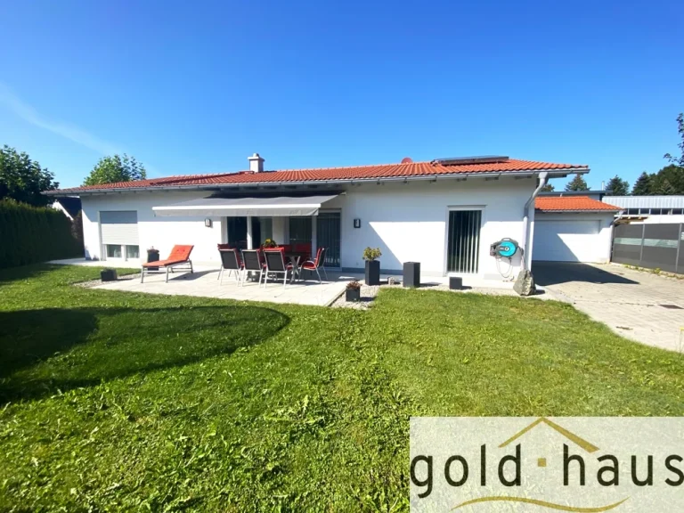 Das Bild zeigt einen modernen Bungalow mit Flachdach, erkennbar durch die rötlichen Dachziegel und die weiße Fassade. Vor dem Haus befindet sich eine gepflasterte Terrasse mit einem Tisch, Stühlen und zwei Liegestühlen. Der Garten ist gepflegt, und es gibt eine Rasenfläche.