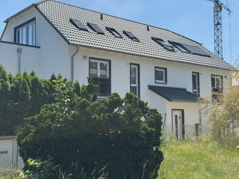 Das Bild zeigt die Eingangsseite eines zweistöckigen Hauses mit Satteldach und Dachfenstern. Die Fassade ist weiß, mit dunklen Fensterrahmen. Vor dem Haus befinden sich dichte Sträucher und ein kleiner, ungepflegter Rasen. Ein Kran ist im Hintergrund sichtbar.