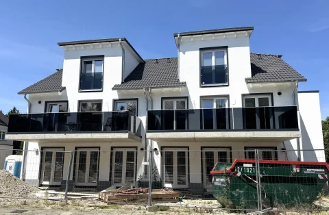 Modernes Doppelhaus in Bau, Gartenseite. Weiße Fassade, schwarze Fensterrahmen, zwei Stockwerke, Satteldächer. Erdgeschoss mit Terrasse und Glasgeländer, 1. Stock mit Balkonen. Baustellenmaterialien und -abfälle im Vordergrund.