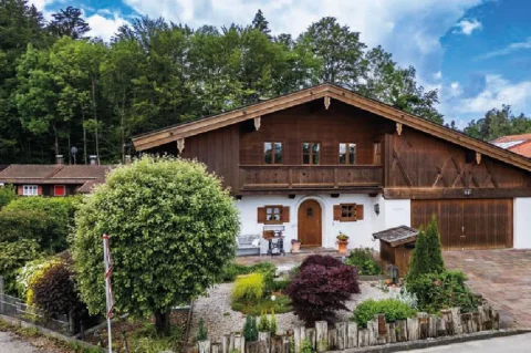 Hausansicht Eingangsbereich Ein typisches, großes alpenländisches Holzhaus in Tegernsee mit verschindelter Fassade, Balkon, weißen Fensterrahmen und einer großen Garage. Vor dem Haus befindet sich ein gepflegter Garten mit Ziersträuchern und einem Kiesweg.