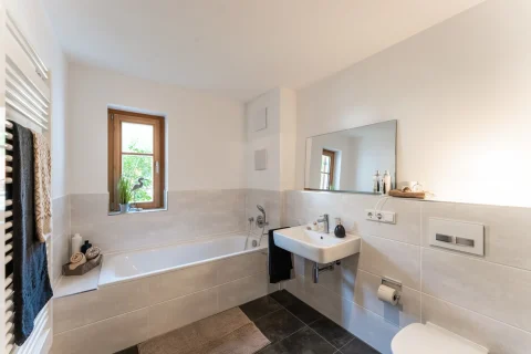 Das Bild zeigt ein helles, modernes Badezimmer in einer Wohnung in Schliersee. Es hat ein Fenster mit Blick auf Grün, eine Badewanne, ein Wand-WC, ein Waschbecken mit Spiegel und eine Wandheizung mit Handtüchern.